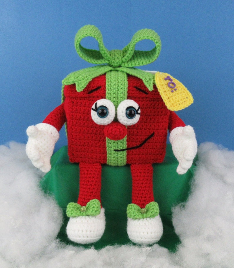 Puede incluir: Un juguete de peluche tejido a crochet con forma de caja de regalo con un lazo verde, un cuerpo rojo y brazos y piernas blancos. El juguete tiene una cara sonriente con ojos grandes y una nariz roja. La caja de regalo tiene una etiqueta verde con la palabra "To!" en ella.