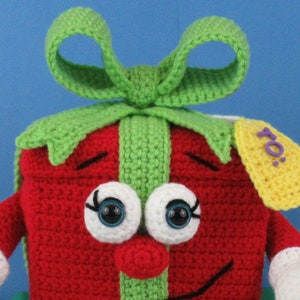 Puede incluir: Una caja de regalo de crochet roja y verde con un gran lazo, ojos blancos con pupilas azules, una nariz roja y una boca sonriente negra. La caja de regalo tiene una etiqueta amarilla con la palabra "Para" escrita en morado.