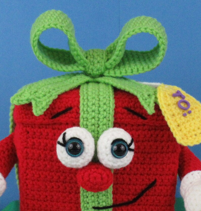 Puede incluir: Una caja de regalo de crochet roja y verde con un gran lazo, ojos blancos con pupilas azules, una nariz roja y una boca sonriente negra. La caja de regalo tiene una etiqueta amarilla con la palabra "Para" escrita en morado.