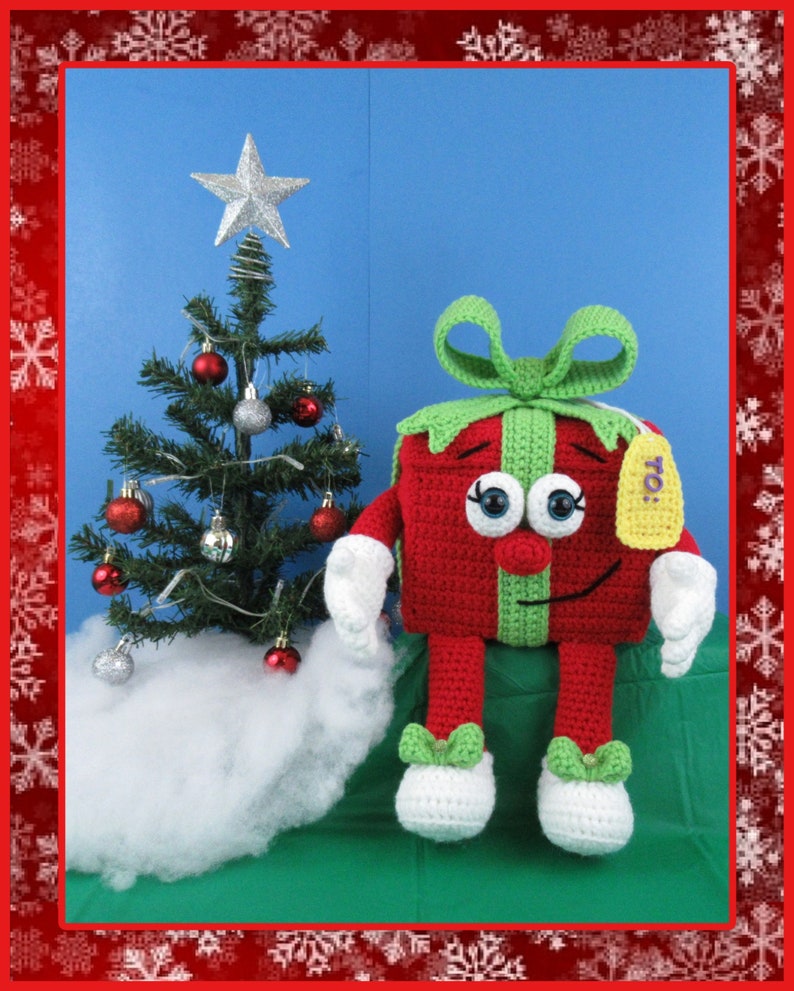 Puede incluir: Una caja de regalo de crochet roja y verde con una etiqueta blanca que dice "To!" est&aacute; sentada frente a un peque&ntilde;o &aacute;rbol de Navidad con adornos rojos y plateados. El &aacute;rbol est&aacute; decorado con luces blancas y est&aacute; sentado sobre una superficie blanca y esponjosa.