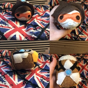 May include: A handmade plush toy of a character from the video game Overwatch. The toy is brown, white, and orange and has a black eye mask. The toy is sitting on a red, white, and blue fabric.