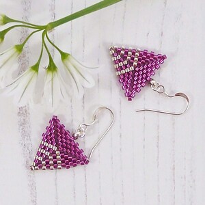 May include: A pair of magenta and silver beaded triangle earrings with silver hooks. The earrings are handmade with a geometric pattern. White flowers and a green stem are in the background, set against a light wooden surface.