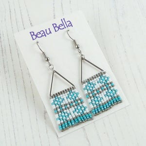 May include: A pair of dangle earrings with a triangular silver frame and beaded fringe. The fringe features a pattern of white, gray, and turquoise beads. The earrings are displayed on a white card with the text "Beau Bella".