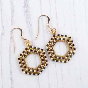 May include: A pair of gold earrings featuring black and white hexagonal bead designs. The earrings are crafted from small, round beads woven together to create a geometric pattern.