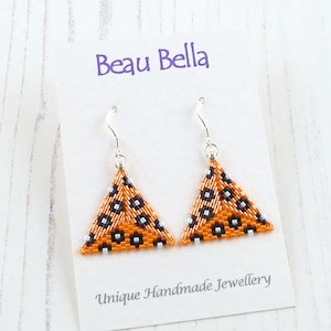 May include: Orange and black triangle-shaped earrings with silver hooks. The earrings are beaded and have a geometric pattern. The earrings are on a white card with the text "Beau Bella" and "Unique Handmade Jewellery".