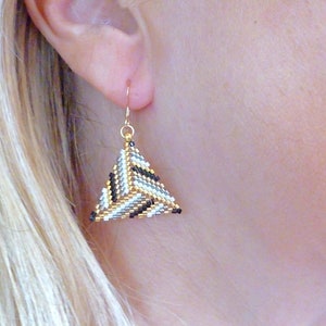 Peut inclure: Boucles d'oreilles en forme de triangle avec un motif géométrique en perles noires, blanches et dorées.