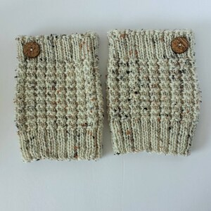 May include: A pair of knitted leg warmers in a speckled off-white yarn. Each leg warmer has a ribbed cuff and is adorned with a wooden button. The textured knitwear features flecks of black, brown, and orange.