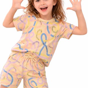 Könnte beinhalten: Ein zweiteiliger Kinder-Schlafanzug in Hellgelb mit einem sich wiederholenden Muster aus blauen, rosa und gelben abstrakten Formen und Text. Das Set besteht aus einem kurzärmeligen Oberteil und passenden Shorts.