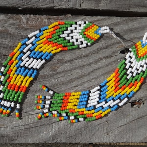 May include: A pair of long, colorful beaded earrings with a geometric pattern. The earrings are made of small, colorful beads in shades of red, orange, yellow, green, blue, white, and black. The earrings are hanging from silver hooks.