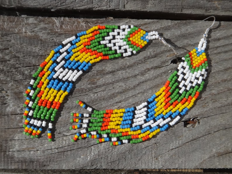 May include: A pair of long, colorful beaded earrings with a geometric pattern. The earrings are made of small, colorful beads in shades of red, orange, yellow, green, blue, white, and black. The earrings are hanging from silver hooks.
