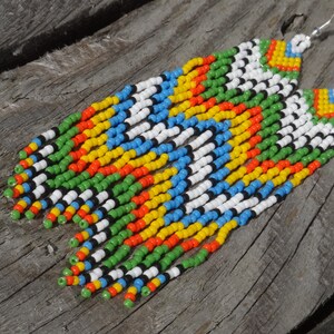 May include: A pair of long, colorful beaded earrings with a geometric pattern. The earrings are made with white, black, green, blue, yellow, and orange beads.