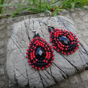 Puede incluir: Un par de pendientes colgantes con cuentas negras y rojas y una piedra preciosa negra en el centro. Los pendientes est&aacute;n sobre una superficie de madera.