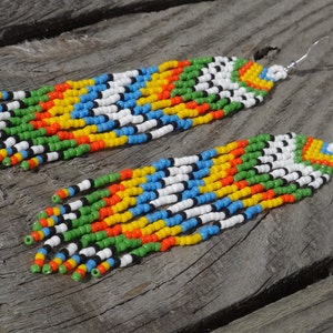 May include: A pair of long, colorful beaded earrings. The earrings are made with small, round beads in shades of green, yellow, orange, blue, white, and black. The beads are strung together in a repeating pattern, creating a striped effect. The earrings are attached to silver hooks.