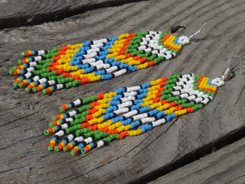 May include: A pair of long, colorful beaded earrings. The earrings are made with small, round beads in shades of green, yellow, orange, blue, white, and black. The beads are strung together in a repeating pattern, creating a striped effect. The earrings are attached to silver hooks.