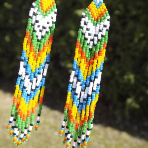 May include: A pair of long, colorful beaded earrings. The earrings are made with small, colorful beads in a variety of colors, including white, black, green, blue, yellow, and orange. The beads are strung together in a pattern that creates a geometric design.