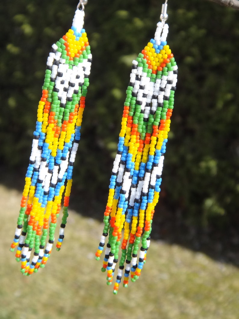 May include: A pair of long, colorful beaded earrings. The earrings are made with small, colorful beads in a variety of colors, including white, black, green, blue, yellow, and orange. The beads are strung together in a pattern that creates a geometric design.