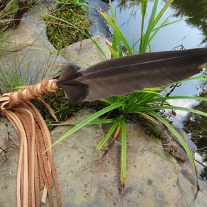 May include: A handcrafted smudge stick with a large black feather, wrapped in tan leather, and adorned with long leather fringe. The smudge stick rests on a gray rock near water and green plants. This item is used for spiritual cleansing and smudging.
