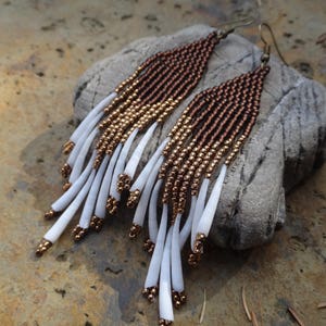 Peut inclure: Une paire de longues boucles d'oreilles en perles avec un dégradé de couleur brun et or. Les boucles d'oreilles ont une frange blanche en bas.