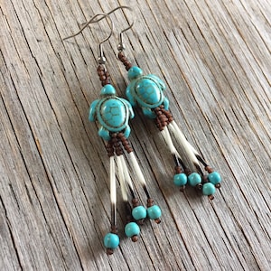 Peut inclure: Boucles d'oreilles tortue turquoise avec des détails marron et blancs. Elles présentent des pendentifs en forme de tortue turquoise, des perles marron et des brins blancs ressemblant à des piquants, pour un design unique inspiré de la nature.