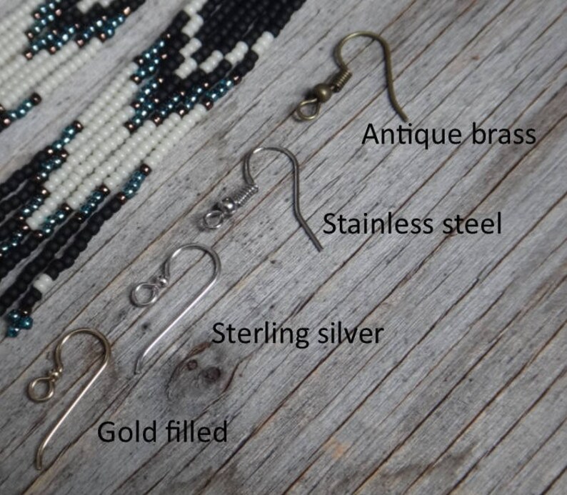 May include: Four different earring hooks are shown on a wooden surface. The hooks are labeled "Antique brass", "Stainless steel", "Sterling silver", and "Gold filled".