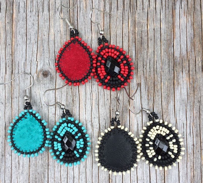 Puede incluir: Tres pares de pendientes de cuentas con detalles de cuero. Los pendientes son de color rojo, turquesa y negro con cuentas negras y una piedra preciosa negra en el centro.