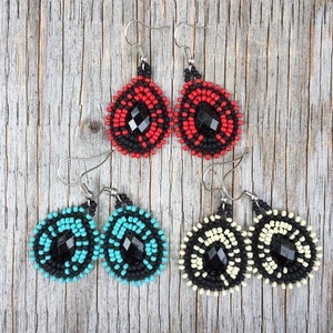 Puede incluir: Tres pares de pendientes de cuentas con centros de piedras preciosas negras. Los pendientes son de color rojo, turquesa y crema.