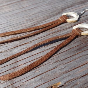Può includere: Un paio di orecchini con nappe in pelle marrone e anelli in osso bianco. Gli orecchini sono appesi a ganci d'argento.