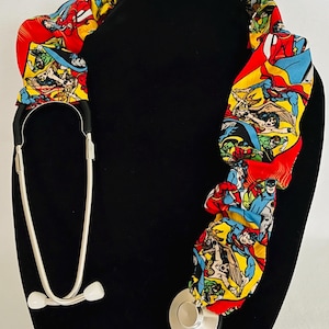 May include: A stethoscope with a red, yellow, and blue fabric cover featuring a comic book pattern of superheroes. The fabric cover is designed to wrap around the stethoscope.