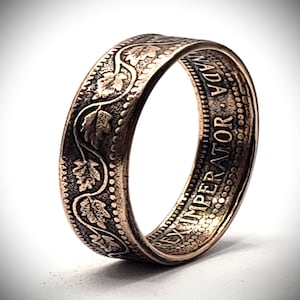 May include: A metallic ring crafted from a coin, featuring a raised vine and leaf pattern on the outer band. The inner surface displays the embossed text CANADA and REX IMPERATOR, with a beaded border detail along the edge of the band.