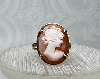 May include: A gold ring with a cameo featuring a white silhouette of a woman's head on a peach-colored background.