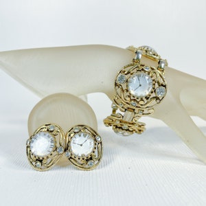 Puede incluir: Un conjunto de joyas vintage con un brazalete dorado y pendientes a juego. El brazalete y los pendientes tienen pequeñas esferas de reloj con números romanos, rodeadas de pedrería transparente. El conjunto se exhibe sobre una superficie blanca.