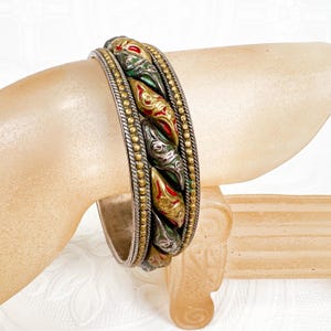May include: A silver-toned bangle bracelet with a central band of green, red, and gold decorative elements. The bracelet has a braided design with a row of small gold beads on either side. The bracelet is on a light-colored surface.