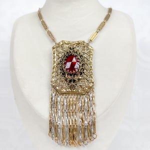 Op de afbeelding: Goudkleurige statement ketting met een grote, gefacetteerde rode edelsteen in het midden. De hanger heeft een open ontwerp en bungelende gouden en zilveren kettingen. De ketting wordt getoond op een witte mannequin.