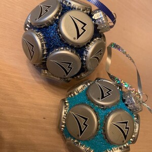 May include: Two Christmas ornaments made from bottle caps. The ornaments are covered in glitter and have a blue and turquoise color scheme. The bottle caps have a silver logo with a black arrow.