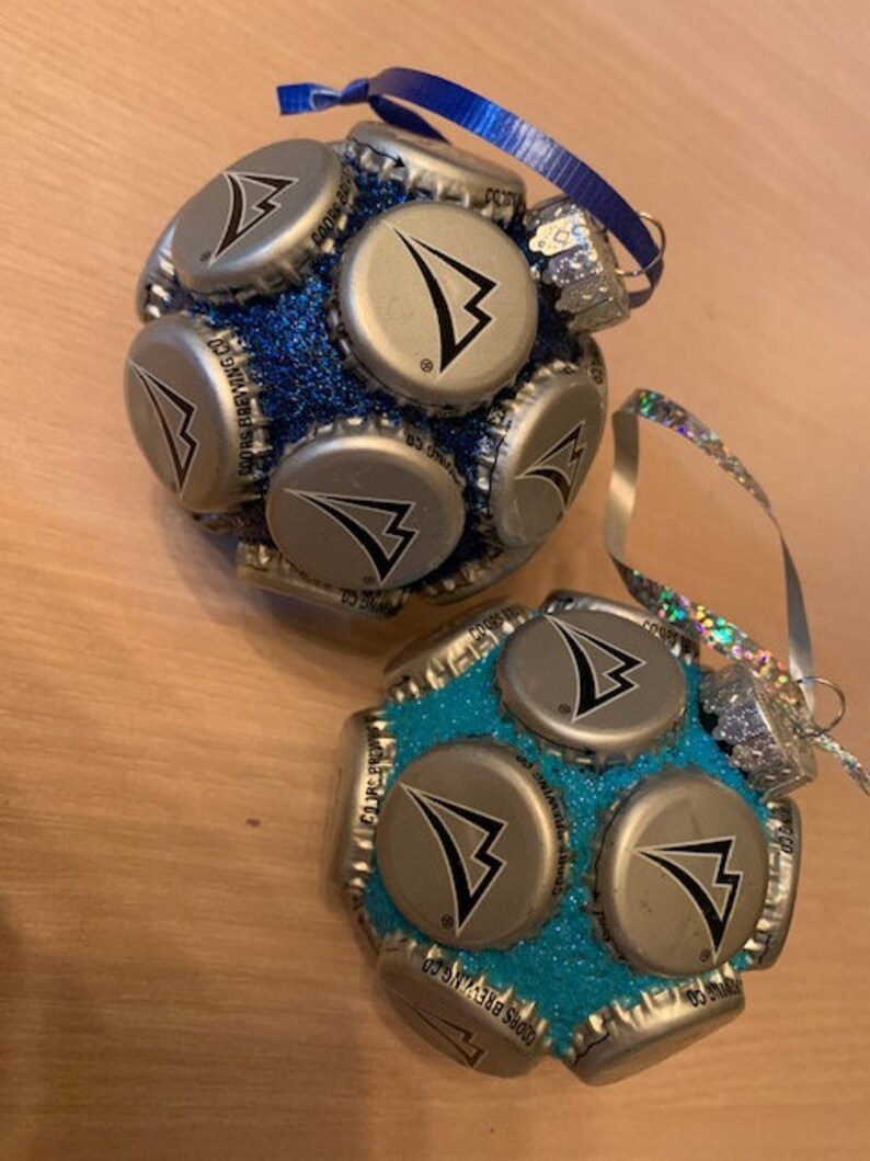 May include: Two Christmas ornaments made from bottle caps. The ornaments are covered in glitter and have a blue and turquoise color scheme. The bottle caps have a silver logo with a black arrow.
