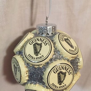 May include: A Christmas ornament shaped like a sphere, made of multiple bottle caps with a silver glitter finish. Each bottle cap features the Guinness logo and the text "Guinness Extra Stout".