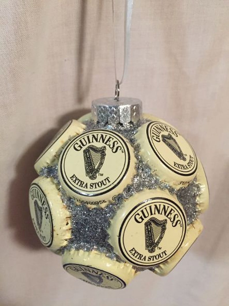 May include: A Christmas ornament shaped like a sphere, made of multiple bottle caps with a silver glitter finish. Each bottle cap features the Guinness logo and the text "Guinness Extra Stout".
