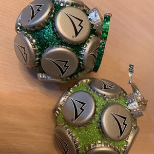 May include: Two green and silver Christmas ornaments made from bottle caps. The ornaments are covered in green glitter and have a silver cap with a black triangle logo.
