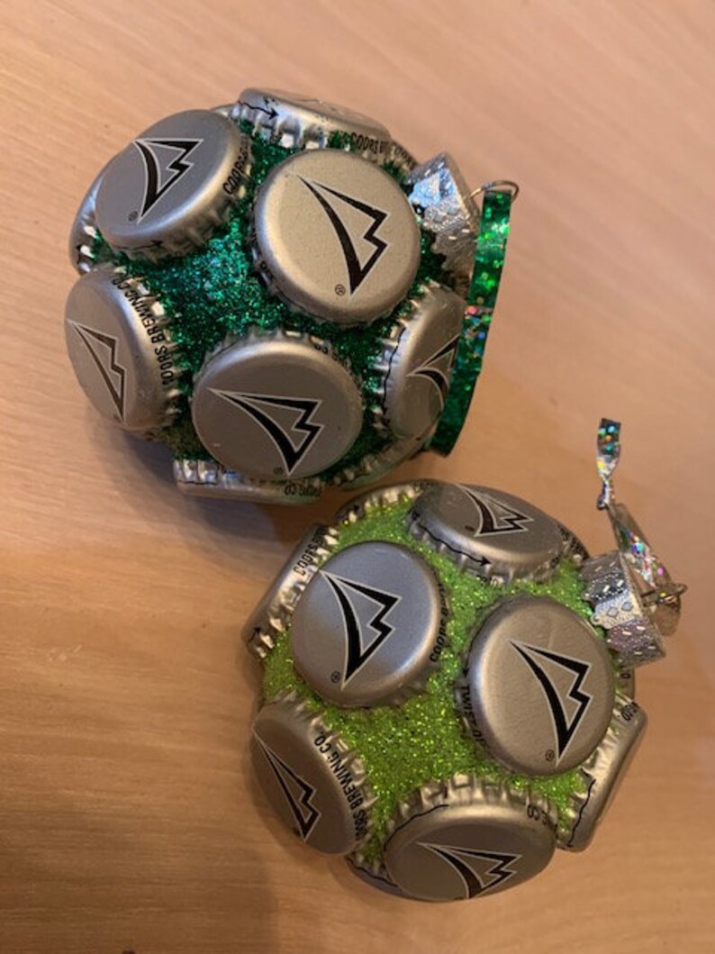May include: Two green and silver Christmas ornaments made from bottle caps. The ornaments are covered in green glitter and have a silver cap with a black triangle logo.