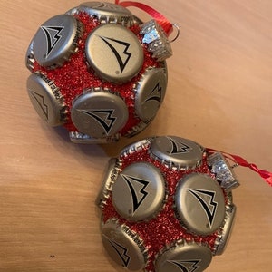 May include: Two red and silver Christmas ornaments made from bottle caps. Each ornament is covered in red glitter and features a silver bottle cap with a black arrow logo.