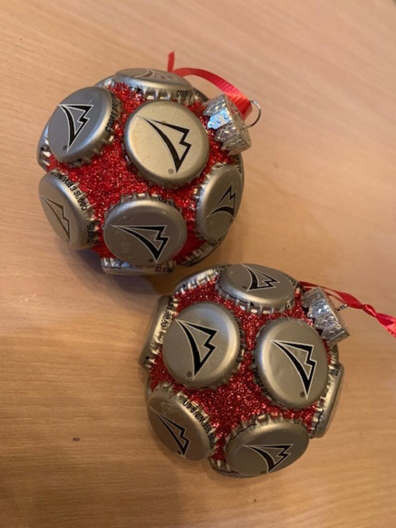 May include: Two red and silver Christmas ornaments made from bottle caps. Each ornament is covered in red glitter and features a silver bottle cap with a black arrow logo.