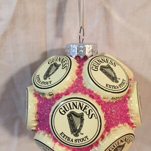 May include: A Christmas ornament made from multiple Guinness beer bottle caps, glued together and covered in pink glitter. The caps feature the Guinness logo and the words "Extra Stout".