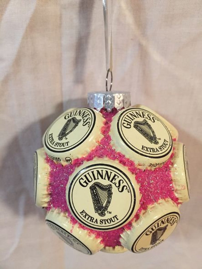 May include: A Christmas ornament made from multiple Guinness beer bottle caps, glued together and covered in pink glitter. The caps feature the Guinness logo and the words "Extra Stout".