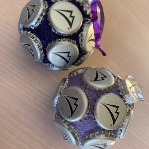 May include: Two purple and silver Christmas ornaments made from bottle caps. The ornaments are decorated with a silver arrow logo and the text "Coors Brewing Co."