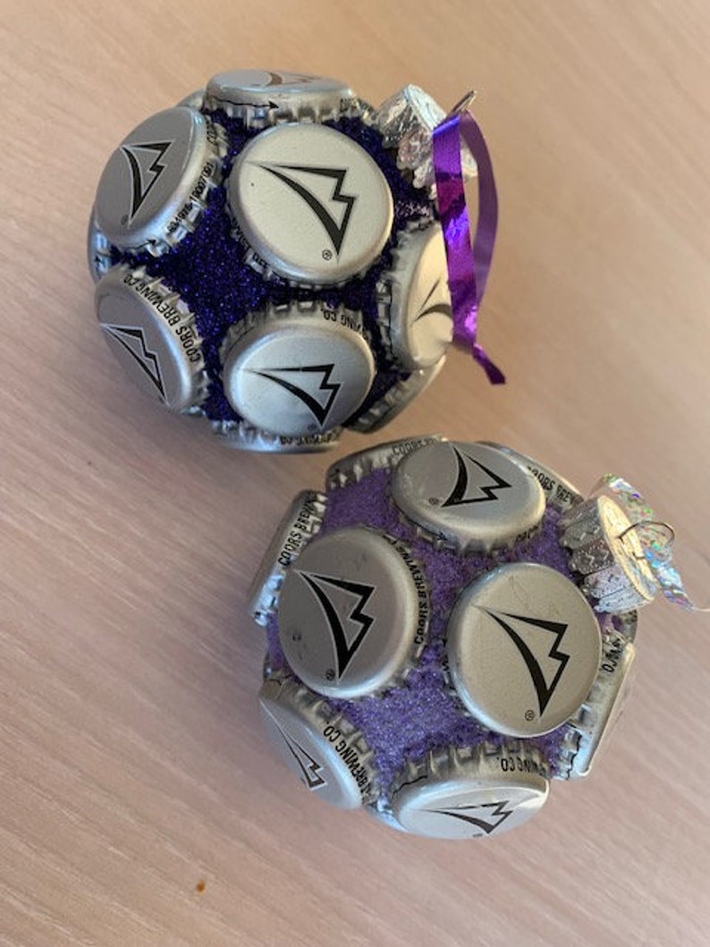 May include: Two purple and silver Christmas ornaments made from bottle caps. The ornaments are decorated with a silver arrow logo and the text "Coors Brewing Co."