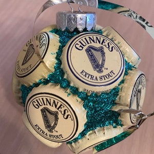May include: A Christmas ornament made from multiple Guinness beer bottle caps, with a green glitter background. The caps are arranged in a circular pattern and feature the Guinness logo and the words "Extra Stout".