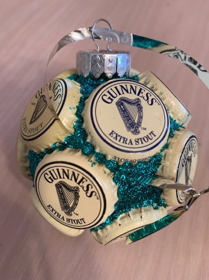 May include: A Christmas ornament made from multiple Guinness beer bottle caps, with a green glitter background. The caps are arranged in a circular pattern and feature the Guinness logo and the words "Extra Stout".