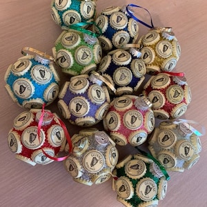 May include: A collection of 14 Christmas ornaments made from Guinness beer bottle caps. Each ornament is a sphere covered in bottle caps and features a different color and glitter finish. The ornaments are decorated with the Guinness logo and harp.