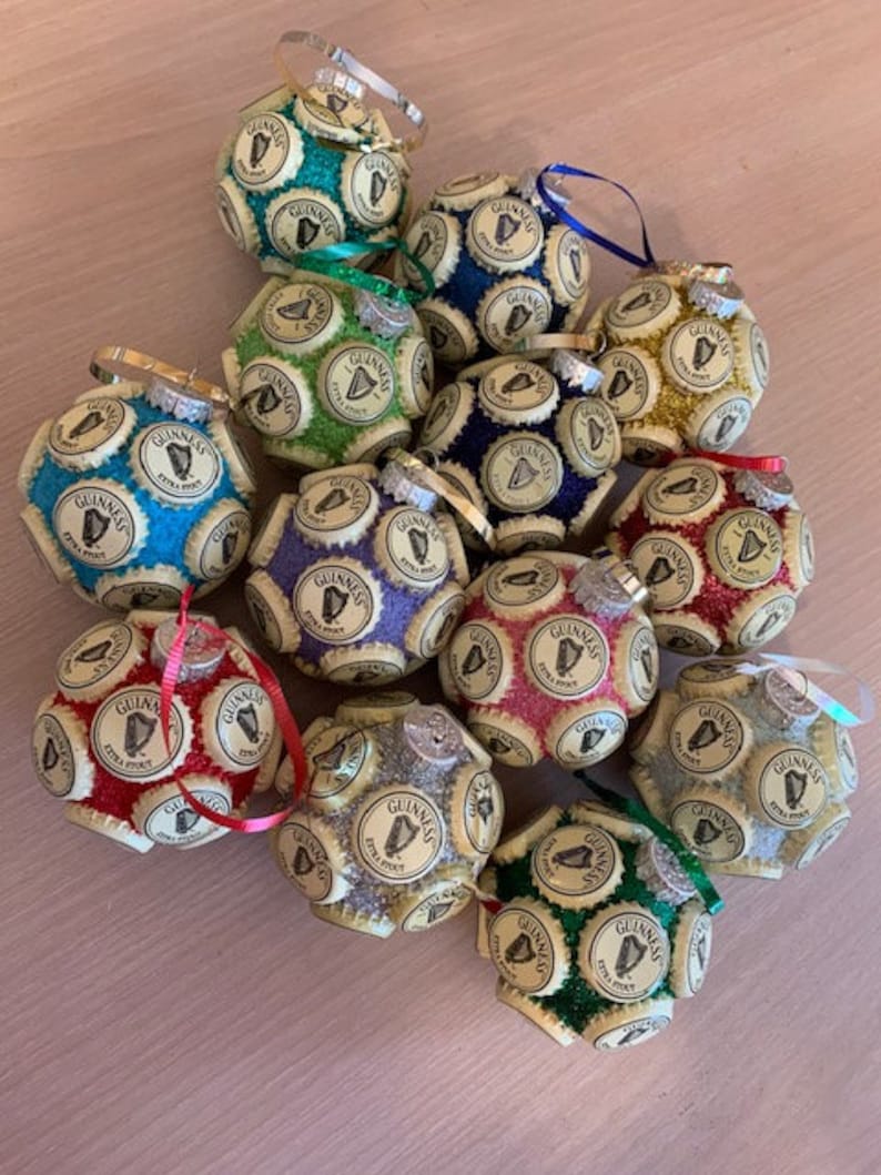 May include: A collection of 14 Christmas ornaments made from Guinness beer bottle caps. Each ornament is a sphere covered in bottle caps and features a different color and glitter finish. The ornaments are decorated with the Guinness logo and harp.