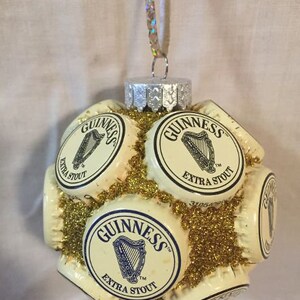 May include: A Christmas ornament made from multiple Guinness beer bottle caps, covered in gold glitter. The caps are arranged in a sphere shape and feature the Guinness logo and the words "Extra Stout".